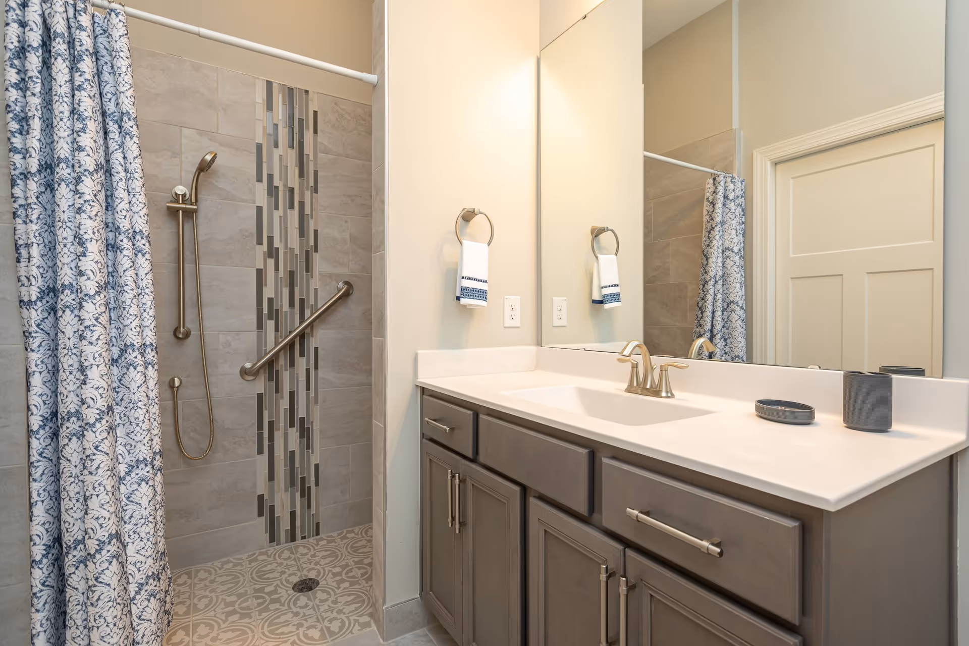 A bathroom featuring a walk-in shower with a handheld showerhead and a grab bar mounted on the tiled wall. The shower has a decorative vertical strip of mosaic tiles and a patterned floor. Next to the shower is a vanity with a white countertop, a built-in sink, and gray cabinets with silver handles. A large mirror is mounted above the vanity, reflecting part of the shower curtain and a closed door. A towel ring with a white hand towel is attached to the wall beside the mirror.