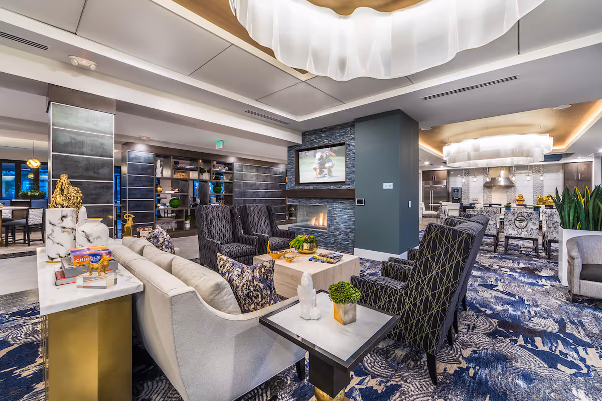 A modern and stylish senior living facility common area featuring a cozy seating arrangement with a sofa and patterned armchairs around a coffee table. A flat-screen TV is mounted above a stone fireplace. The space includes decorative shelving, plants, and a view into a kitchen and dining area with a large chandelier and bar stools. The room has a blue and white patterned carpet and contemporary lighting fixtures.