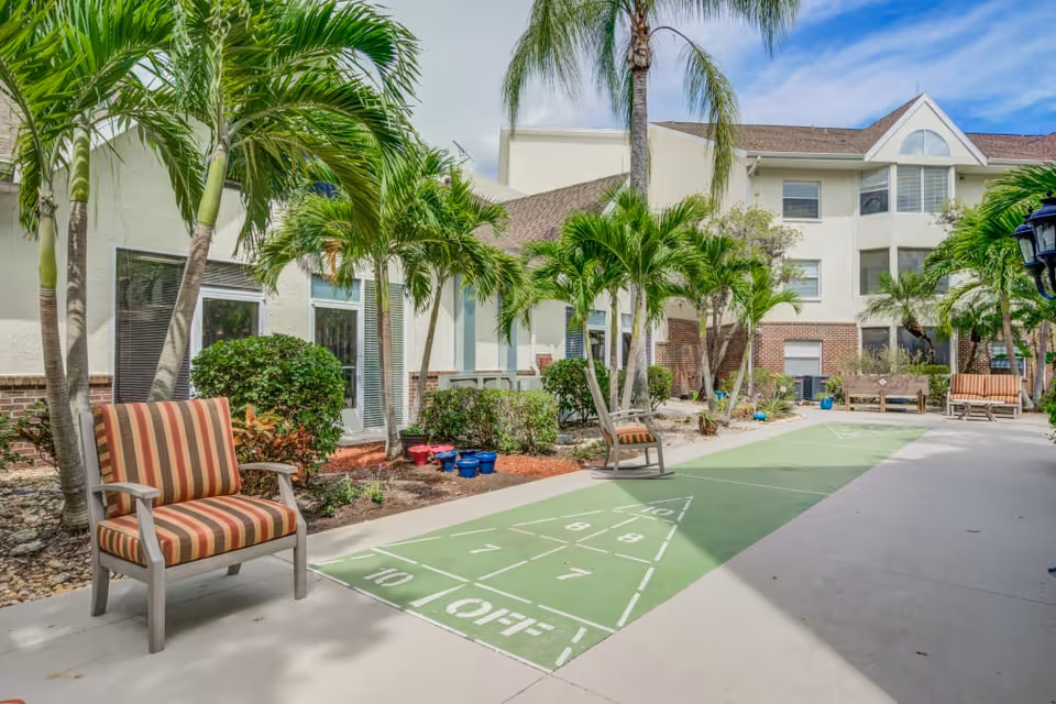 Outdoor courtyard area at Merrill Gardens at Barkley Place featuring a shuffleboard court, several striped cushioned chairs, palm trees, and a multi-story building in the background under a partly cloudy sky.