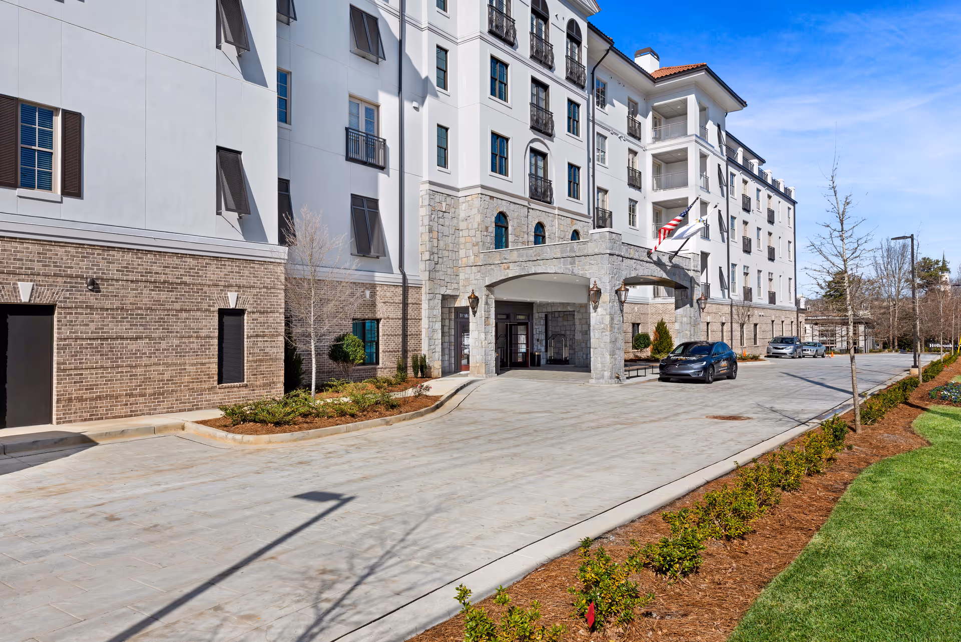 Exterior view of a multi-story senior living facility named Holbrook Decatur with a covered entrance, several windows, and a driveway with parked cars. The building features a combination of brick and stone facade with landscaping along the driveway and a clear blue sky above.