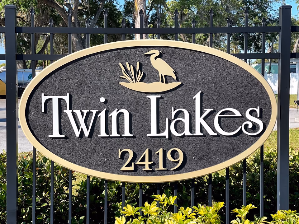 Oval-shaped black and beige sign mounted on a black metal fence with the text 'Twin Lakes 2419' and a graphic of a bird standing on a small patch of land with plants. Green bushes and trees are visible in the background.