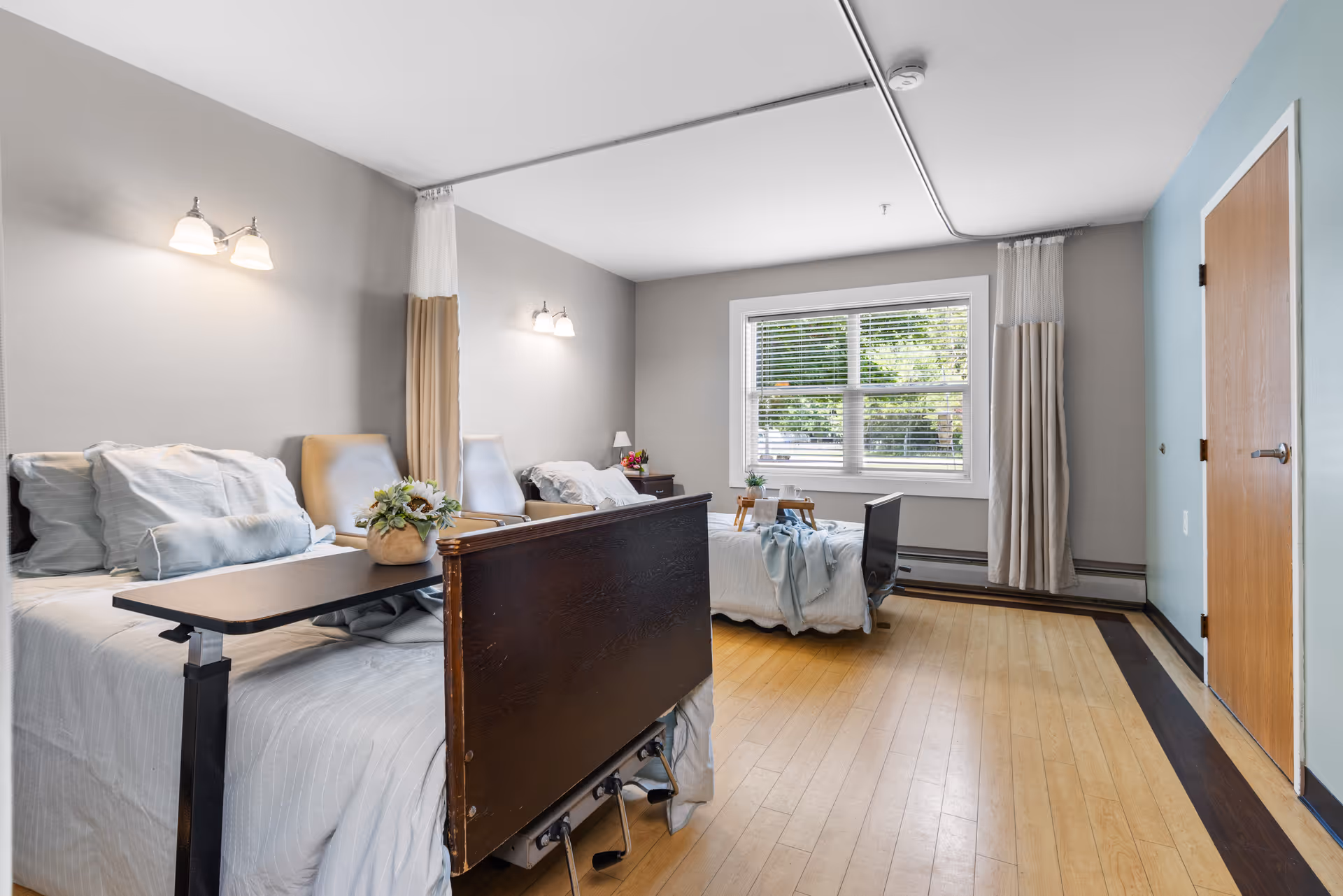 A bright and clean room in a senior living facility with two hospital-style beds, each with white bedding and pillows. There are two beige chairs between the beds, a small table with a flower arrangement, and a large window with blinds letting in natural light. The room has light-colored walls, wooden flooring with a dark border, and a closed wooden door on the right side.