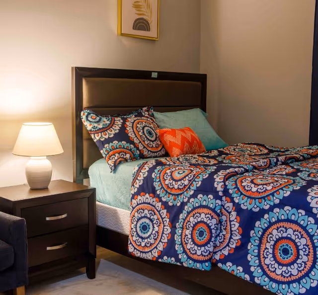 A neatly made bed with colorful patterned bedding and throw pillows next to a nightstand with a lamp and wall art in a cozy bedroom.