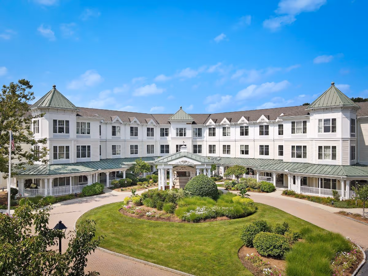 Front exterior of a three-story white senior living building with a circular driveway, landscaped roundabout, and green metal-roofed entrance portico.