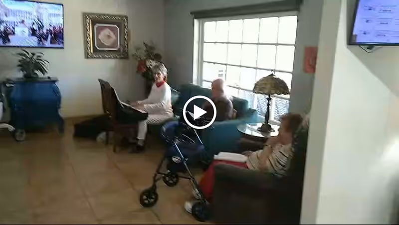 A living room area in a senior care facility with three elderly individuals seated. One woman is playing a keyboard, another person is sitting on a couch, and a third person is seated in a chair with a walker nearby. The room has a large window letting in natural light, a side table with a decorative lamp, a TV mounted on the wall, and framed artwork on the wall.