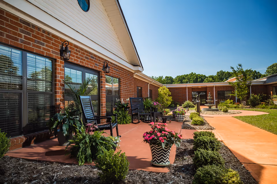 Outdoor patio area at Carolina Care Health and Rehabilitation featuring brick building walls with windows, black chairs, various potted plants and flowers, a walkway, and a fountain in the background under a clear blue sky.