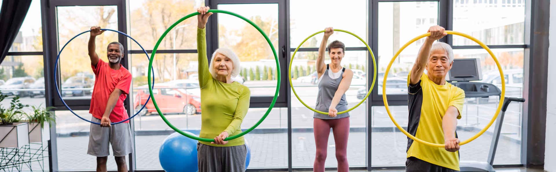 Four seniors standing indoors in front of large windows, each holding a colorful hula hoop above their heads, participating in a group exercise activity with exercise equipment visible in the background.