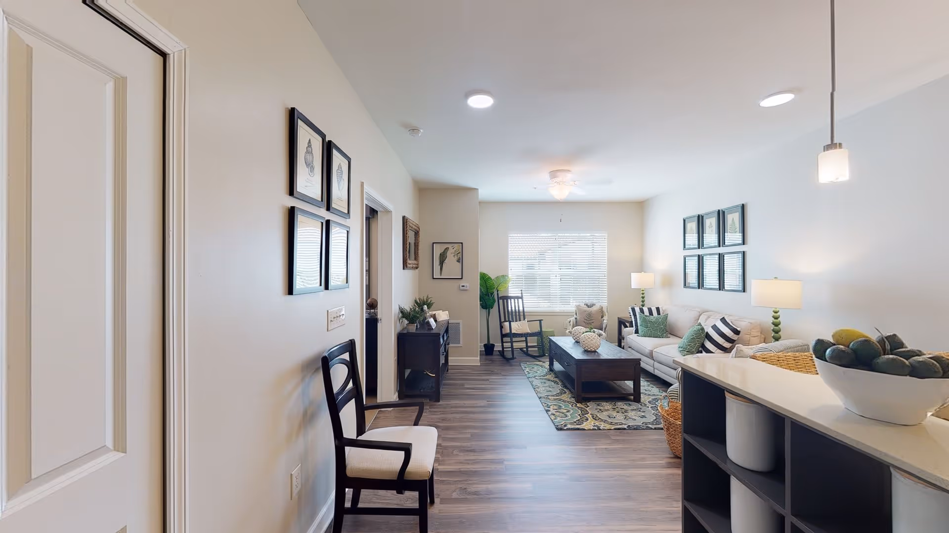 A bright and modern living room with a beige sofa adorned with green and striped pillows, a wooden coffee table on a patterned rug, a rocking chair near a window with blinds, and a small console table with plants and framed pictures on the wall. The room has wood flooring and light-colored walls, with a kitchen counter partially visible on the right side holding a bowl of fruit.