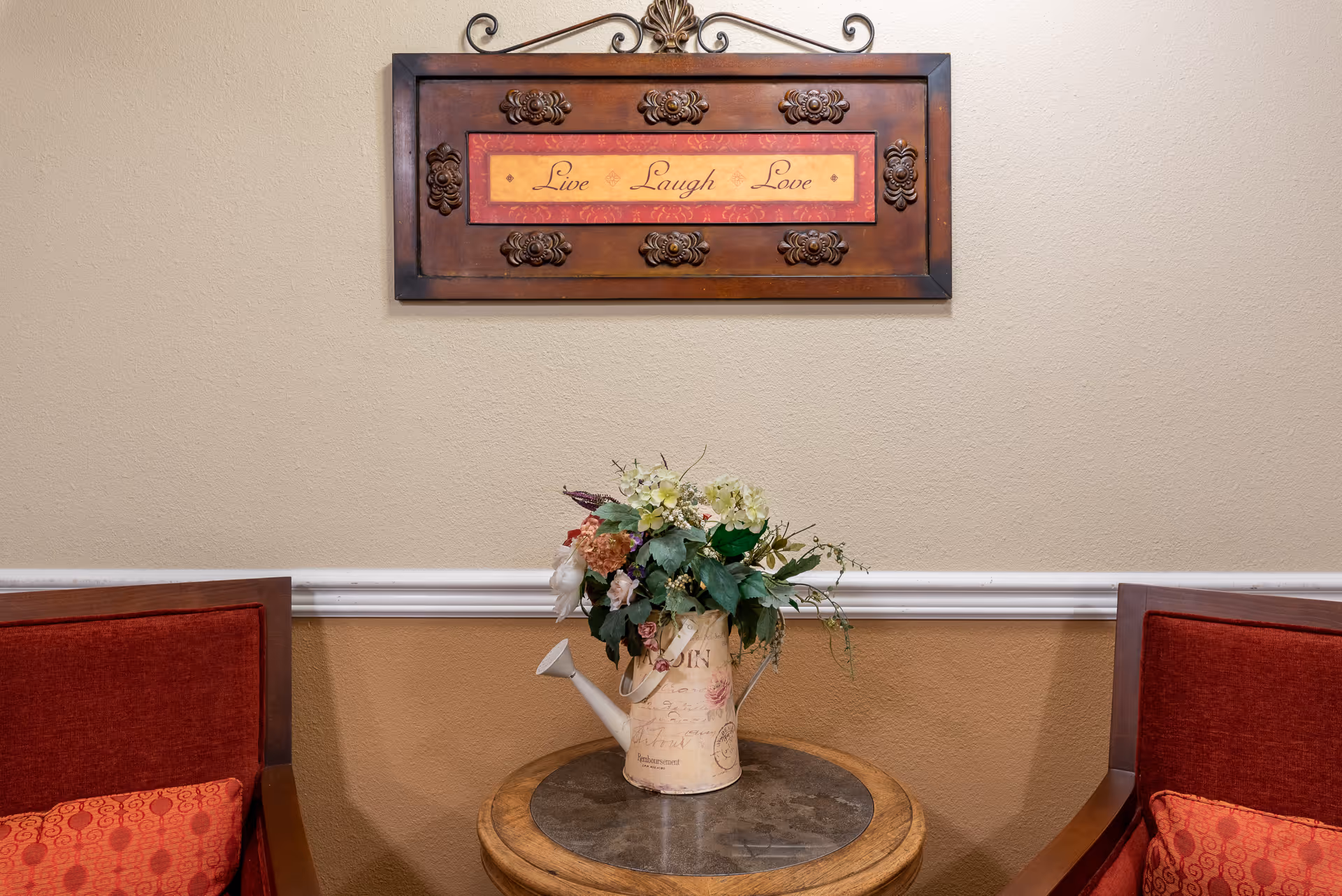 Small seating area with two red chairs flanking a round wooden table holding a floral arrangement in a watering can beneath a framed "Live Laugh Love" wall sign.