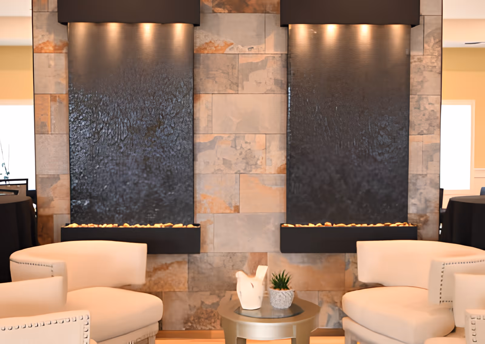 A cozy seating area with two cream-colored armchairs facing each other, separated by a small round table holding a white decorative sculpture and a small potted plant. Behind the chairs are two large vertical water features mounted on a stone wall, each illuminated by three overhead lights.