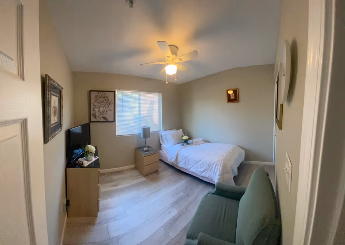 A small, cozy bedroom with a single bed covered in white bedding, a bedside table with a lamp, a small dresser with a TV on top, a green armchair, and framed artwork on the walls. The room has light-colored walls and flooring, a ceiling fan with a light, and a window with blinds.