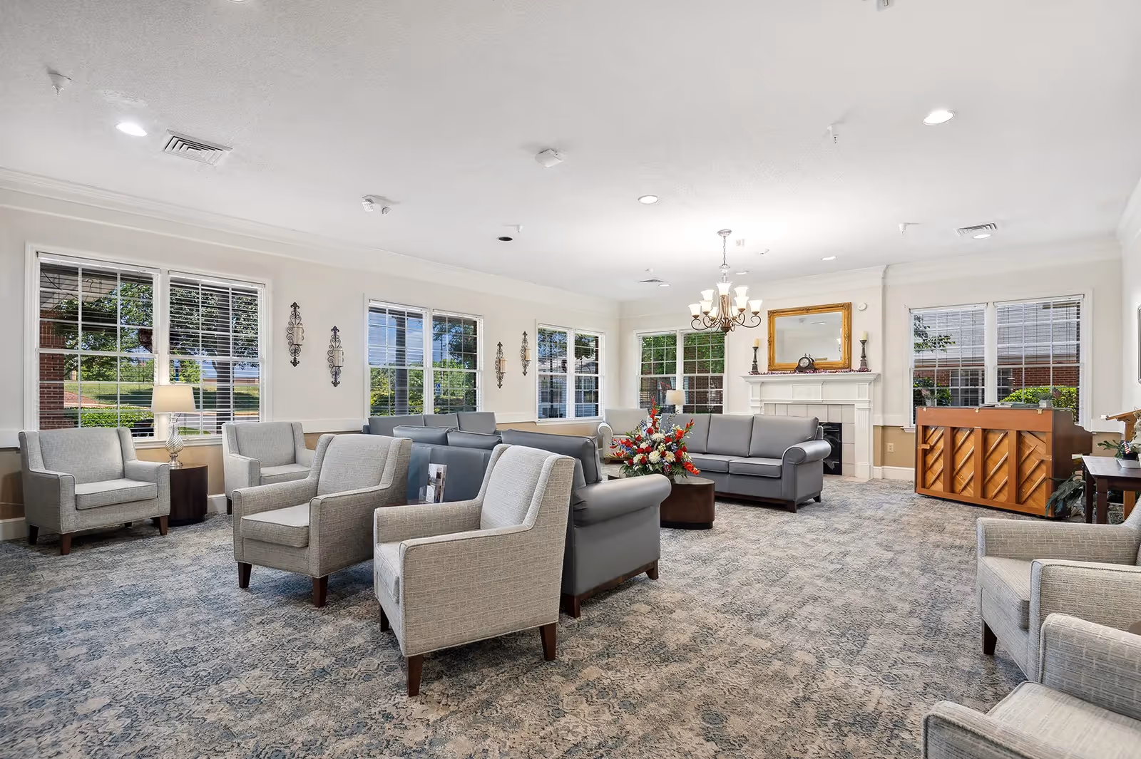 A spacious and well-lit living room with multiple gray armchairs and sofas arranged around a central coffee table with a floral arrangement. The room features large windows with white blinds, a chandelier hanging from the ceiling, a fireplace with a mirror above it, and a wooden piano in the corner. The carpet has a patterned design, and the walls are painted white with decorative sconces.