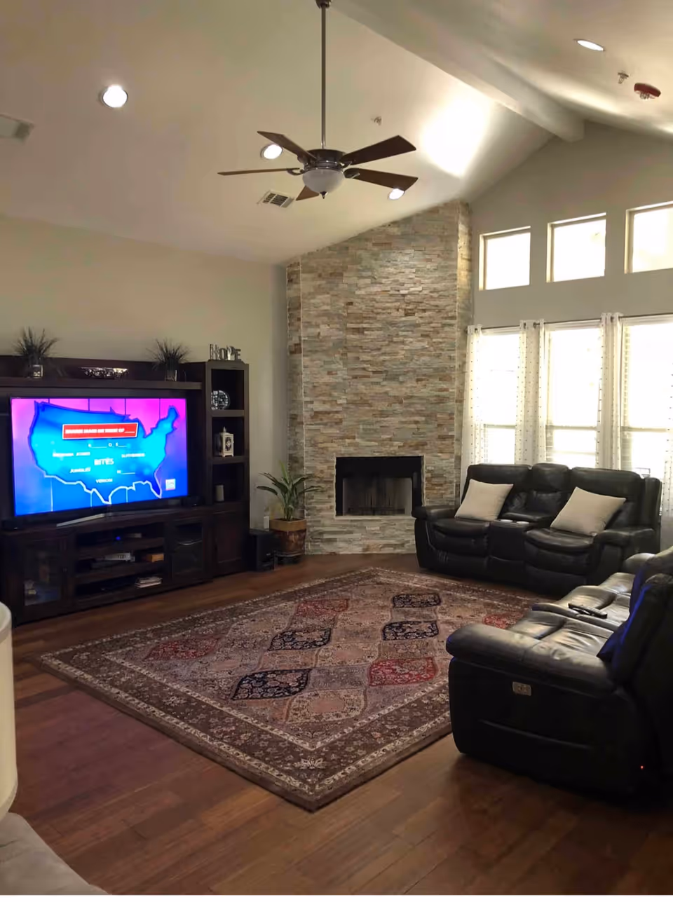 A cozy living room with a large patterned area rug on wooden floors, two black leather sofas with light-colored pillows, a stone fireplace, a ceiling fan, and a dark wooden entertainment center with a flat-screen TV displaying a map of the United States. The room has high ceilings and several windows with sheer curtains allowing natural light to enter.