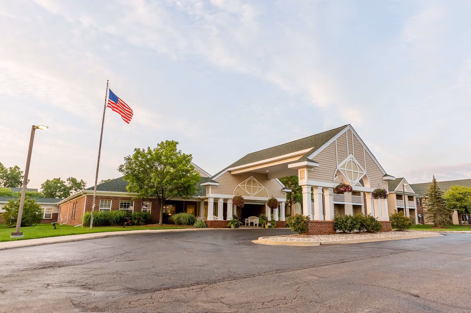 Exterior view of Independence Village of Grand Ledge senior living facility showing a large covered entrance with columns, hanging flower baskets, an American flag on a flagpole, and surrounding greenery under a partly cloudy sky.