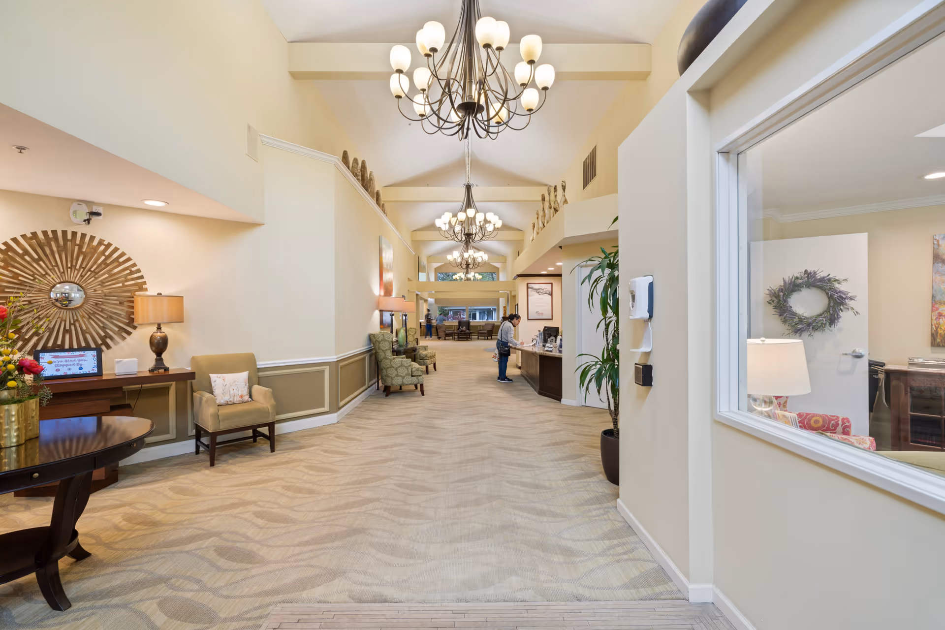 A spacious, well-lit hallway in a senior living facility with beige walls and carpeted floors. The hallway features multiple chandeliers hanging from a vaulted ceiling, several armchairs along the walls, decorative wall art, and a reception desk where a person is standing. A window on the right shows a cozy room with a lamp, wreath, and furniture.
