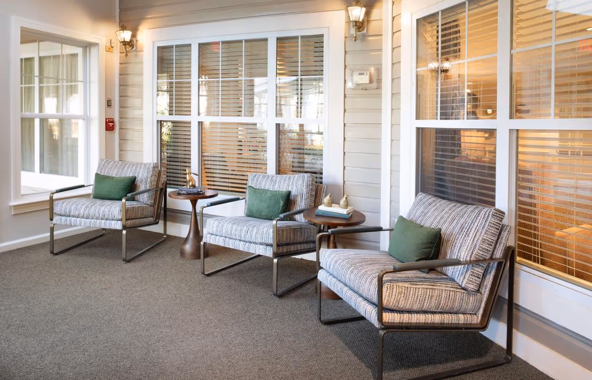 A bright indoor seating area with three upholstered armchairs with green pillows and small round side tables in front of large windows with blinds.