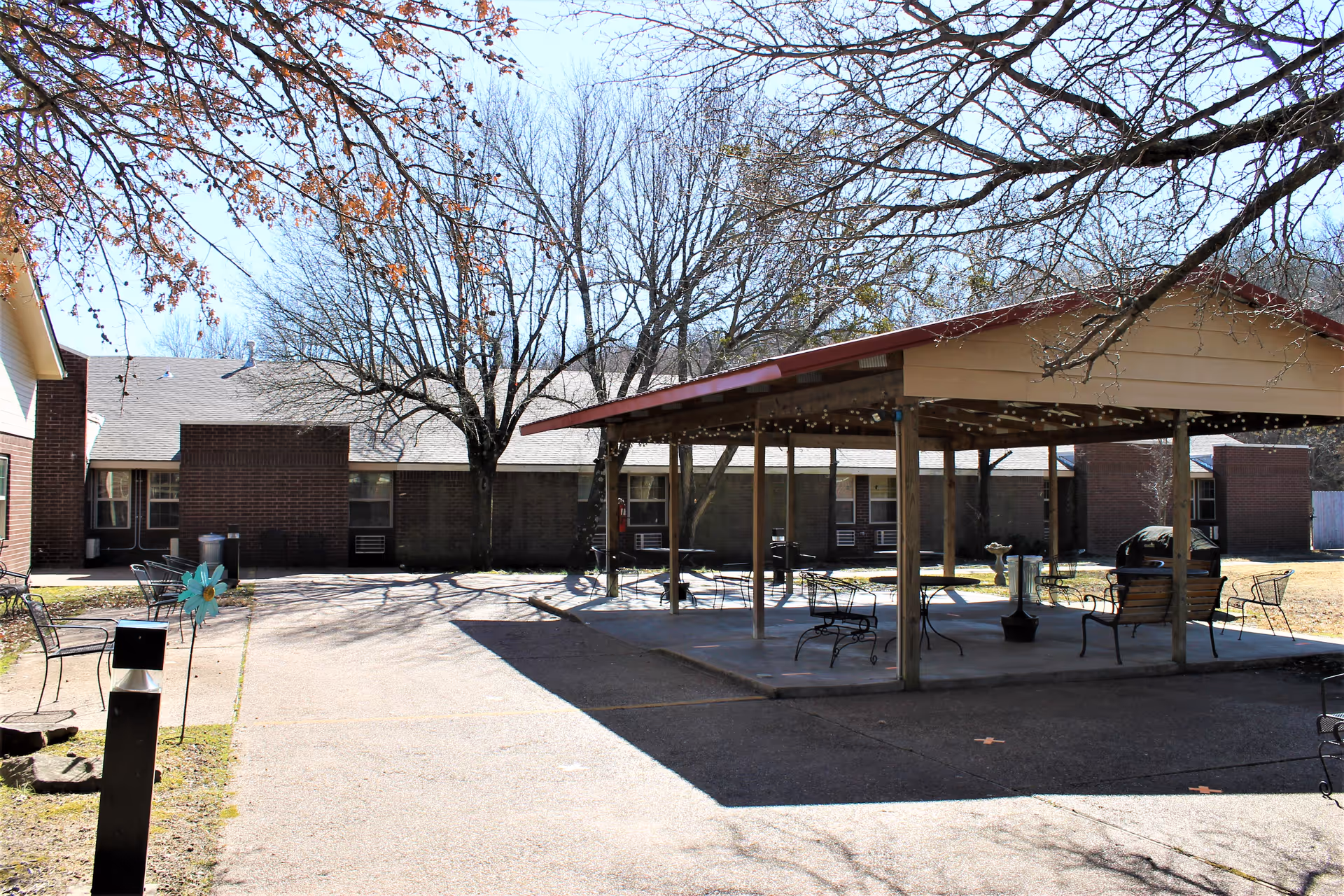Outdoor courtyard area of a nursing home with a covered pavilion featuring benches and chairs. Leafless trees surround the area, and a brick building is visible in the background under a clear blue sky.