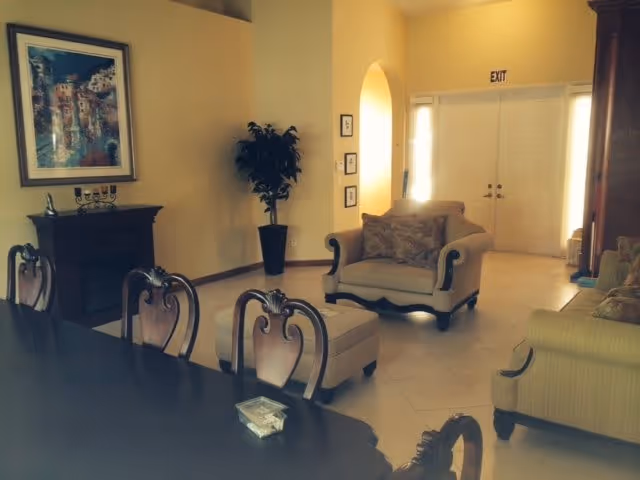 A cozy living room area with beige walls and tiled floor. The room features a beige armchair with a matching ottoman, a beige sofa, a dark wooden dining table with ornate chairs, a potted plant in the corner, a framed painting on the wall, and a double-door exit with an exit sign above it.