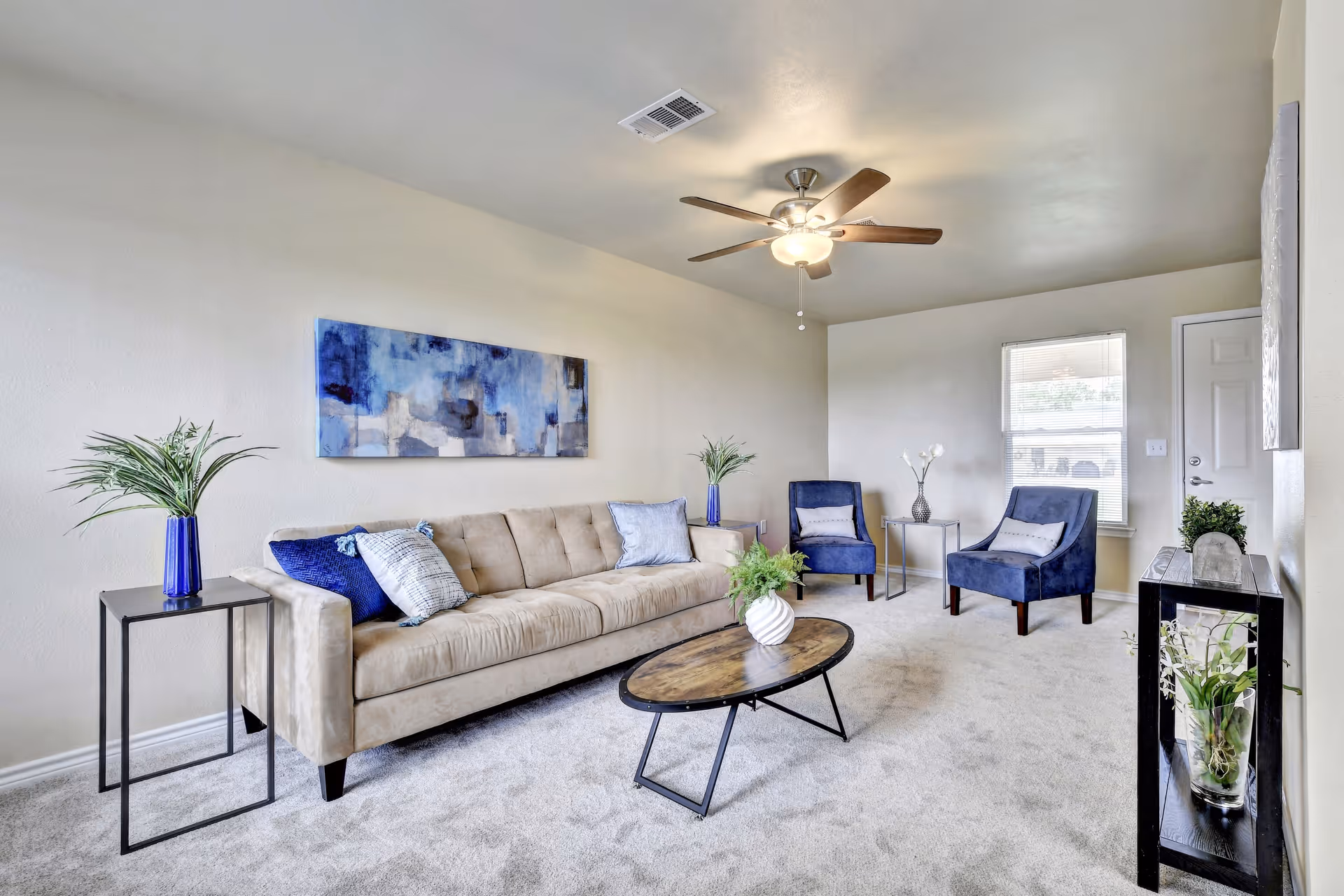 Bright furnished living room with a beige sofa, two blue armchairs, coffee table, ceiling fan, and decorative plants.