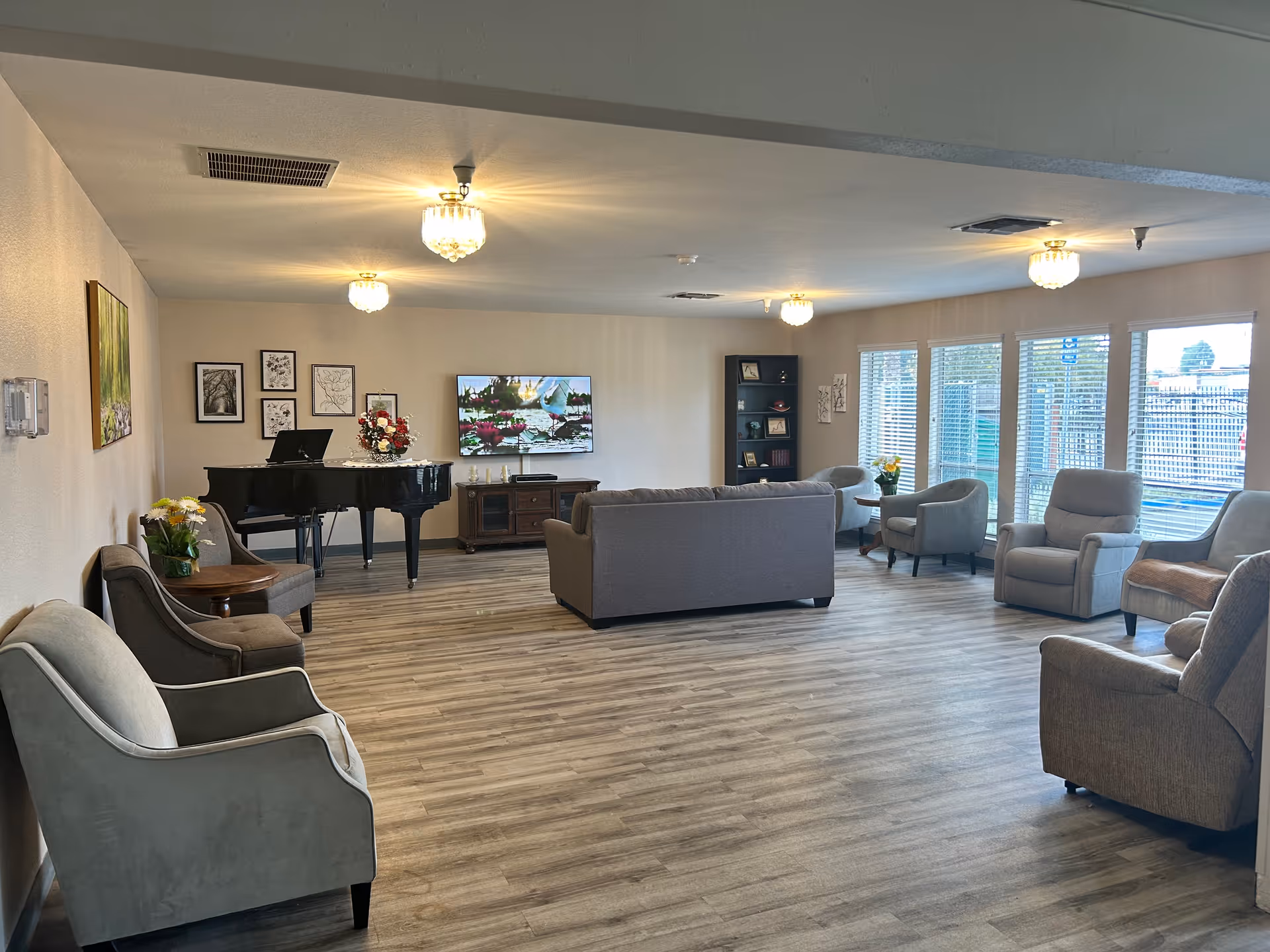 A spacious living room with multiple armchairs and sofas arranged around the room. There is a black grand piano with a floral arrangement on top, a TV mounted on the wall displaying a nature scene, and a bookshelf with decorative items. Large windows with blinds allow natural light to fill the room, and the floor is covered with light wood laminate.