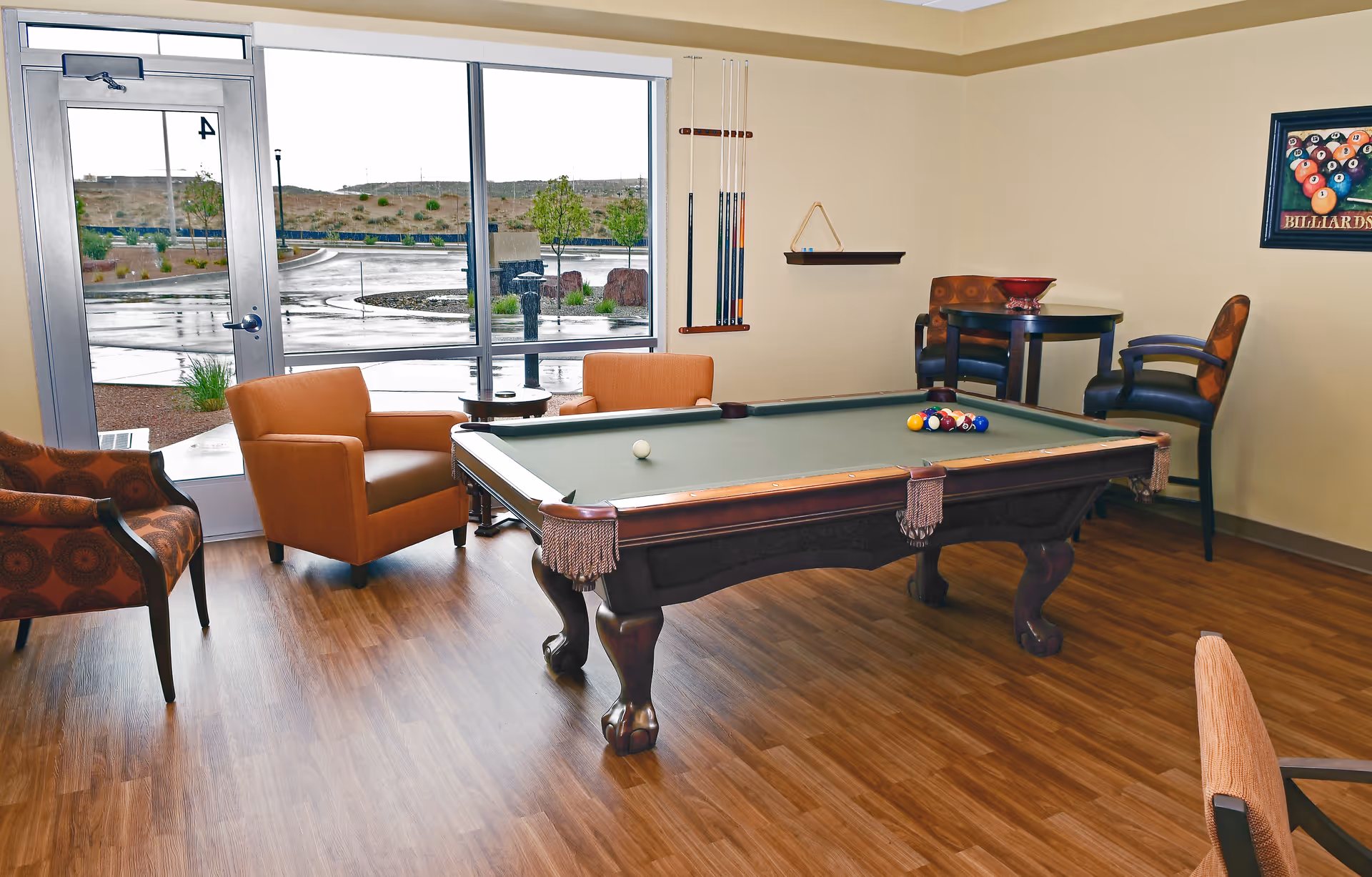 A recreational room with a pool table in the center, surrounded by several chairs and a small table. The room has large windows and a glass door showing an outdoor view with a parking lot and some greenery. Pool cues are mounted on the wall, and a framed picture of billiard balls hangs on the wall.
