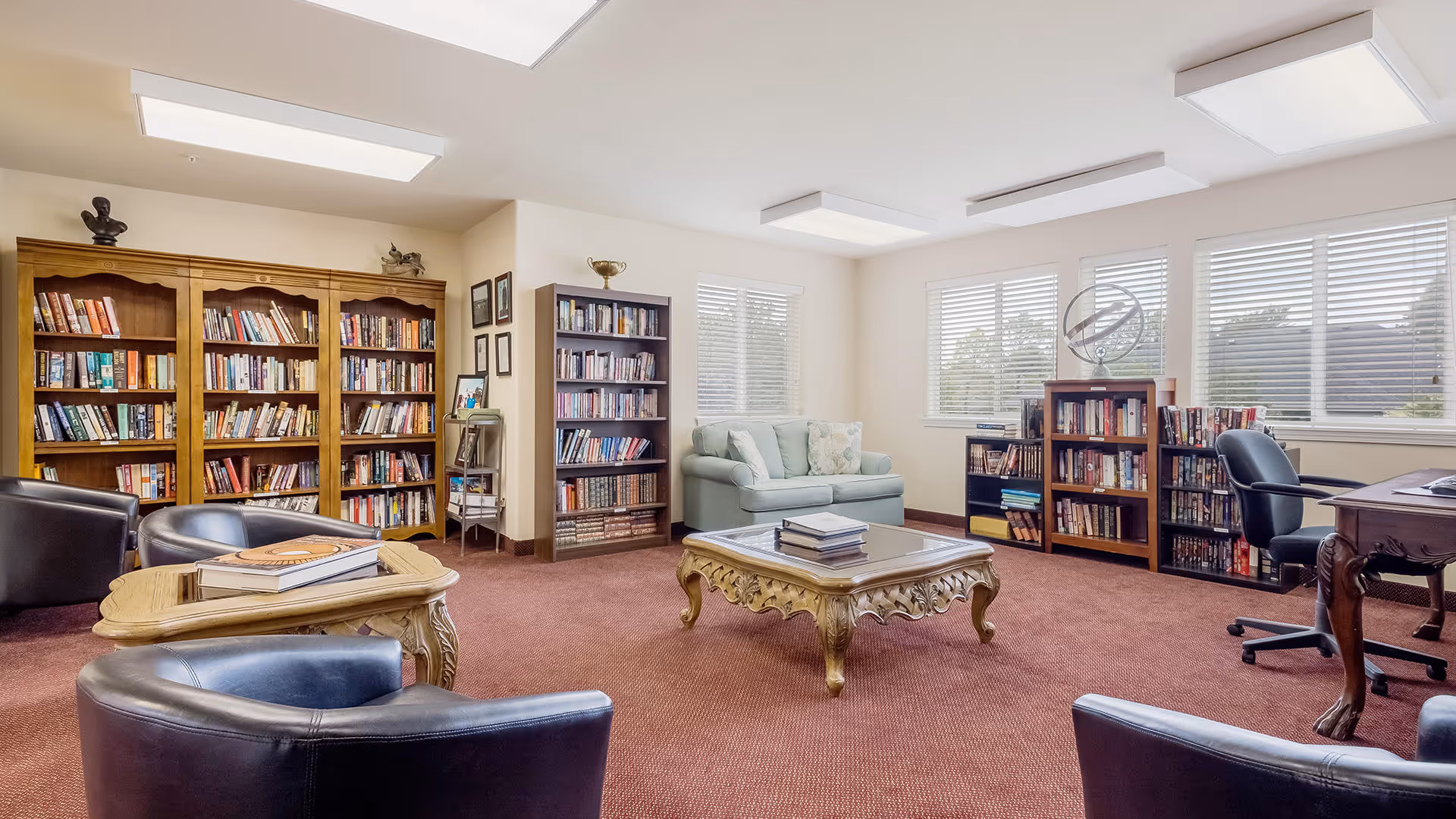 A bright and spacious library room with multiple bookshelves filled with books, a light blue loveseat, several black leather chairs, a wooden coffee table with books on it, and a wooden desk with an office chair near windows with white blinds.