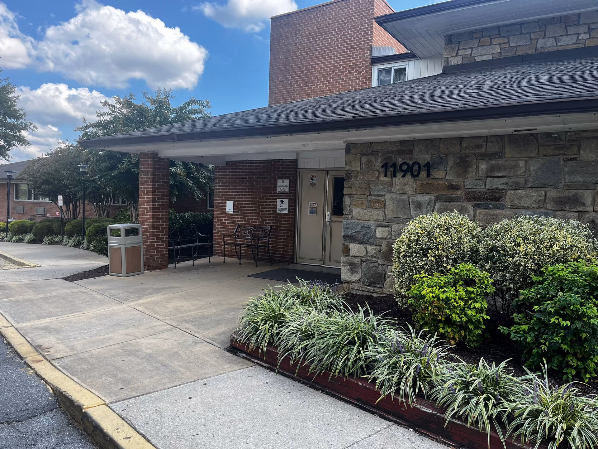 Entrance to a building with a stone and brick exterior, featuring the number 11901 on the wall. There are bushes and plants along the walkway, a covered porch area with benches, and a trash can near the entrance. The sky is partly cloudy.