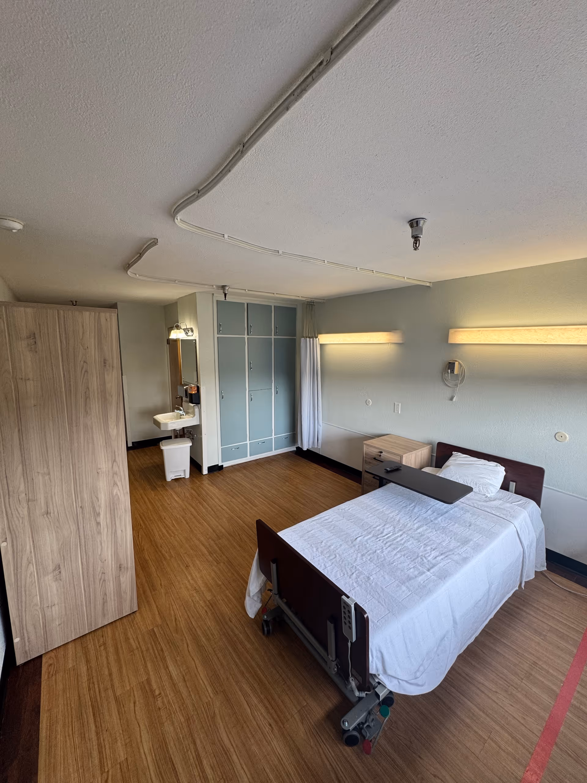 A clean senior care room with a single adjustable bed, bedside table, sink, wardrobe and cabinets on wood flooring.
