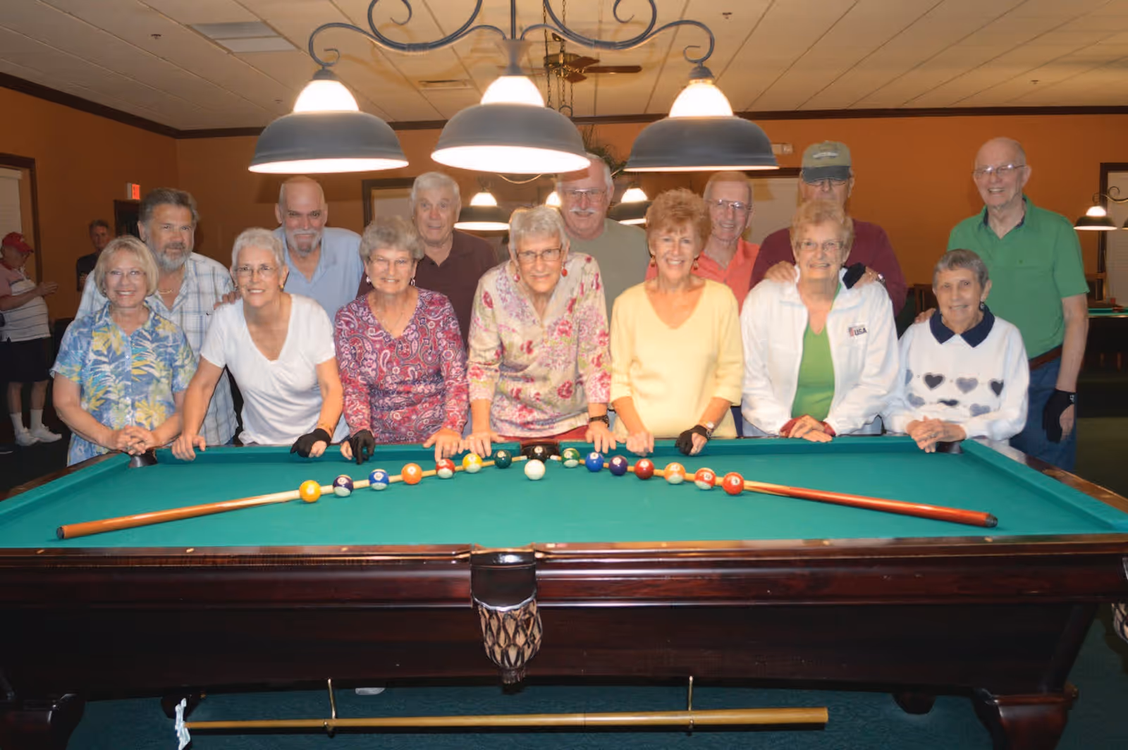 A group of elderly people standing behind a pool table with billiard balls arranged in a line and two pool cues placed on the table. The room is warmly lit with hanging lamps above the table.