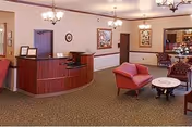 A reception area in a senior living facility with a wooden front desk, two red upholstered chairs, a round marble-top table, framed artwork on the walls, and carpeted flooring. The space is well-lit with ceiling lights and has a warm, welcoming atmosphere.