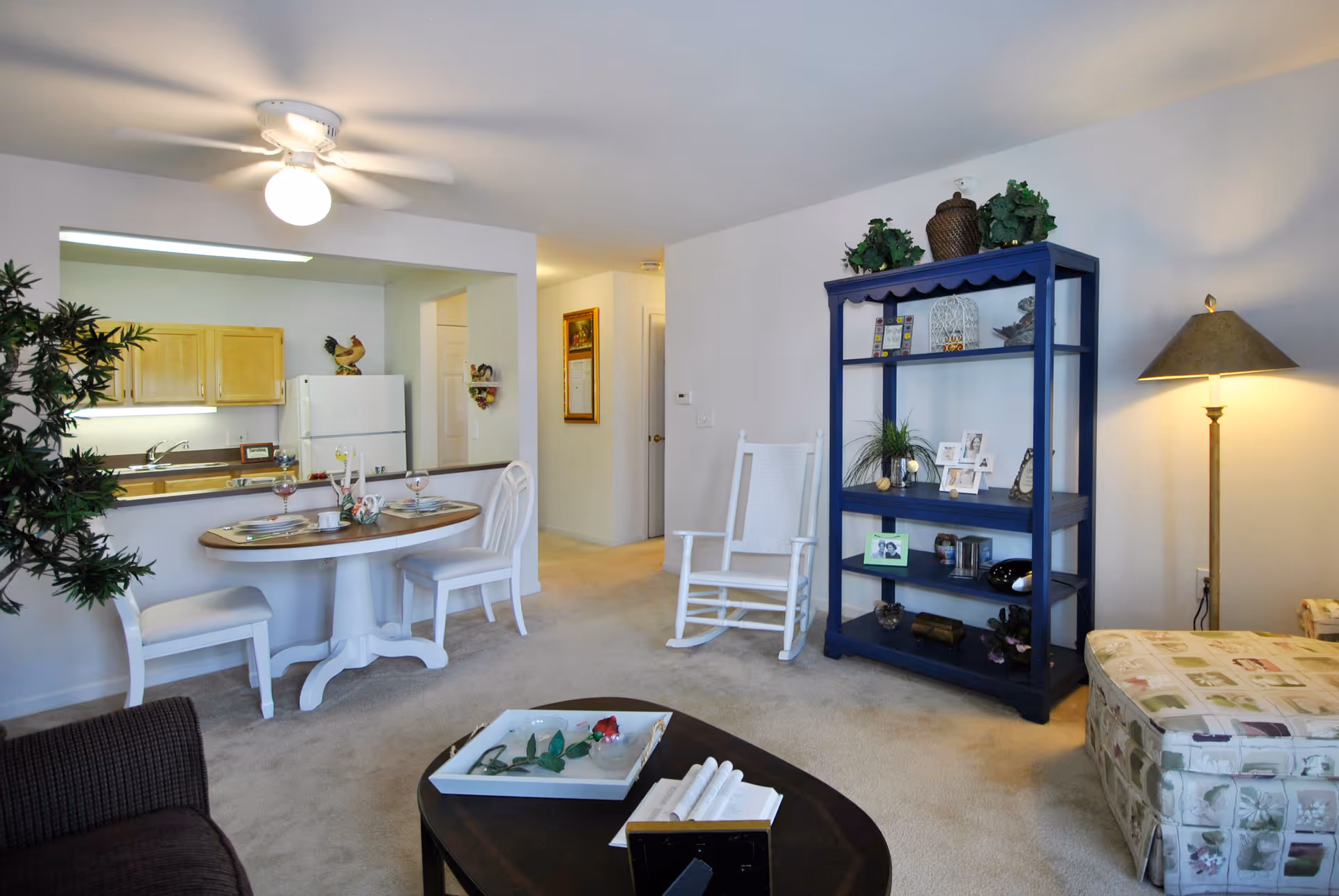 Interior view of a retirement community apartment showing a living room and dining area. The living room has a dark coffee table with a tray and books, a patterned ottoman, a tall blue shelving unit with decorative items and photos, a floor lamp, and a white rocking chair. The dining area has a small round table set for two with white chairs. The kitchen is visible in the background with wooden cabinets and a white refrigerator. The room is carpeted and well-lit with a ceiling fan light.
