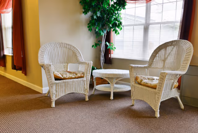 Two white wicker chairs with cushions and a matching wicker coffee table set on a carpeted floor near a large window with blinds and red curtains, next to a tall green potted plant.