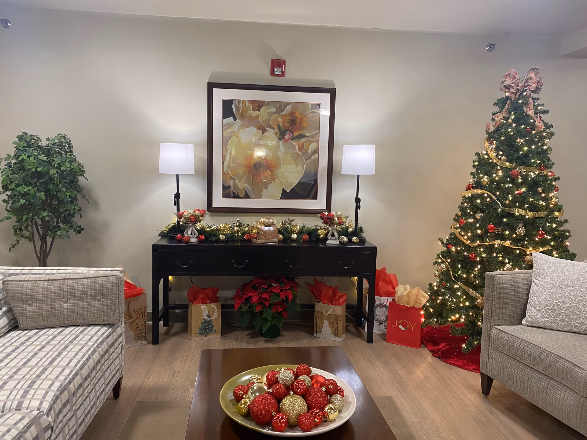 Holiday-decorated living room with a lit Christmas tree, a console table with lamps and ornaments, sofas, and a coffee table bowl of baubles.