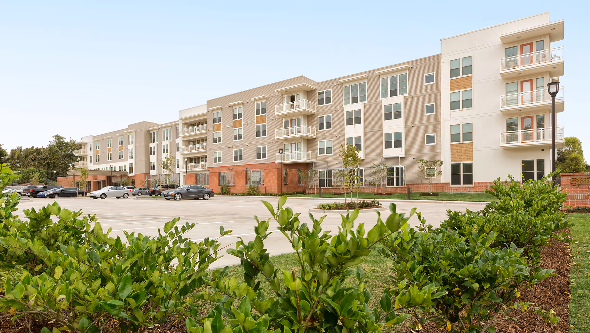 Exterior view of a modern four-story senior living facility building with balconies, large windows, and a parking lot in front. Green shrubs and trees are visible in the foreground under a clear sky.
