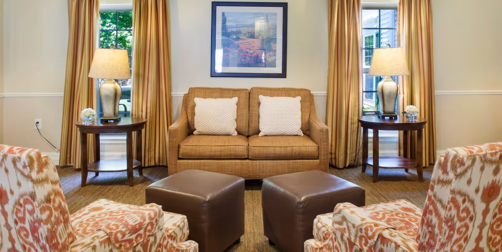 A cozy living room with a brown two-seater sofa with two white pillows, two patterned armchairs, and two brown ottomans. There are two side tables with lamps and small flower arrangements, positioned in front of windows with yellow curtains. A framed picture hangs on the wall above the sofa.