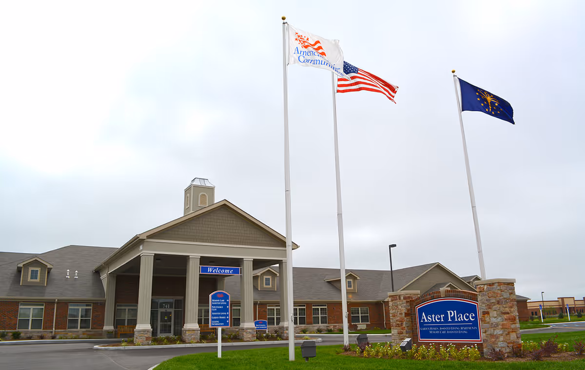 Exterior view of Aster Place senior living facility with a large entrance featuring columns and a covered porch. Three flagpoles display flags including the American flag. A stone sign in front reads 'Aster Place' with additional text about the facility's services. The sky is overcast.
