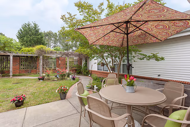 Outdoor patio area with a round table surrounded by six chairs, some with green cushions, and a large patterned umbrella providing shade. The patio is adjacent to a white building with brick trim, and there are potted plants and flowers placed around the concrete walkway. In the background, there is a wooden pergola and a garden area with trees and shrubs.
