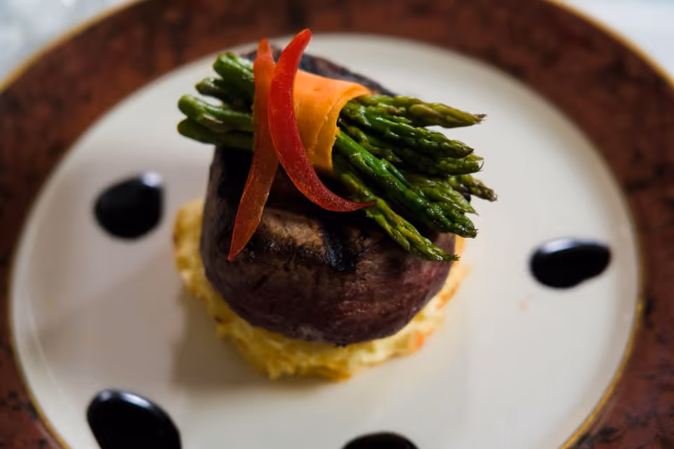 A gourmet dish featuring a grilled filet mignon topped with a bundle of asparagus wrapped in a thin slice of carrot and garnished with red pepper strips, served on a bed of mashed potatoes with decorative black sauce dots on a white plate with a brown rim.
