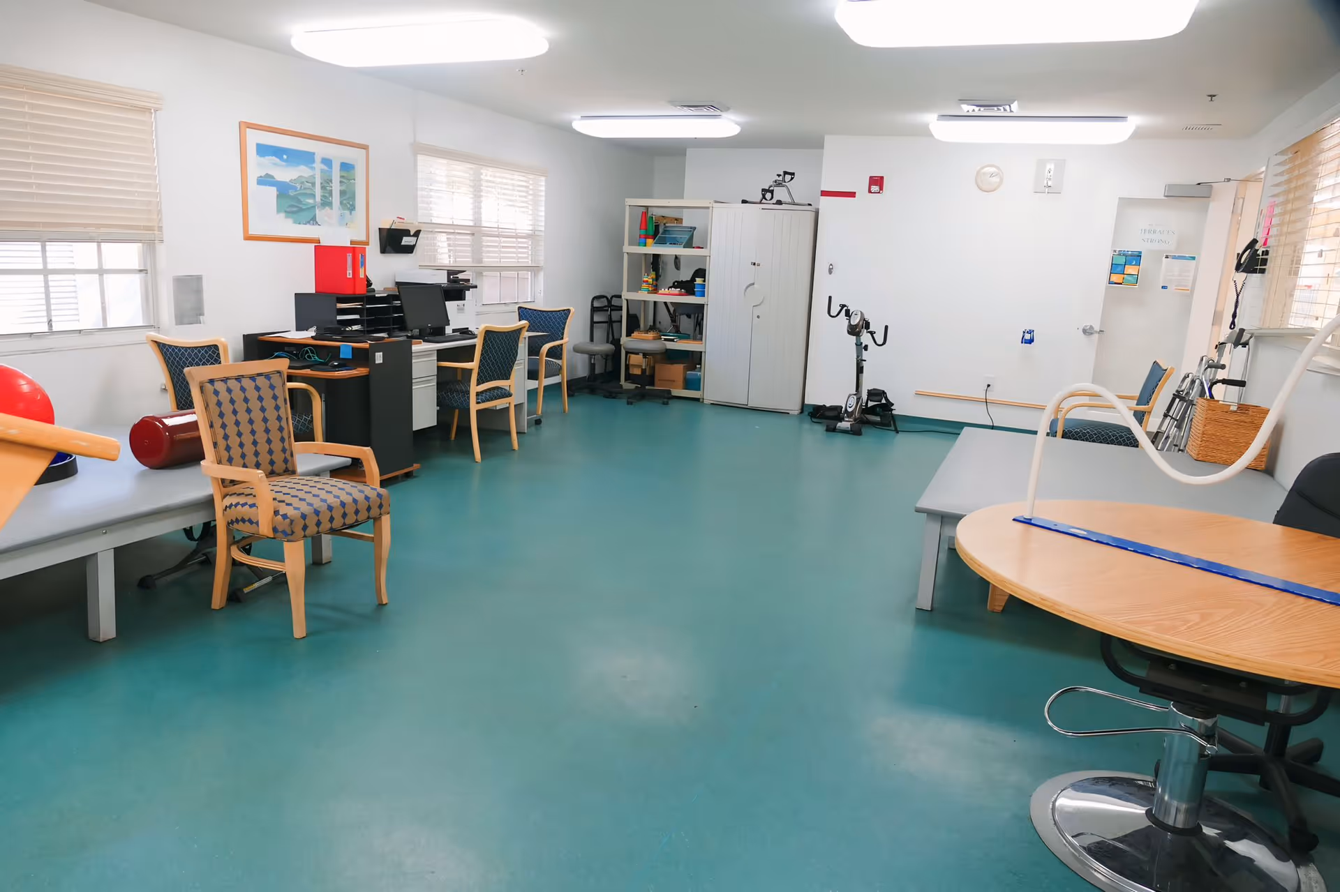 Bright therapy/activity room with chairs, desks, exercise equipment, and therapy tables in a senior care facility.
