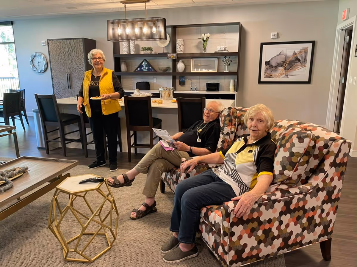 Three residents socialize in a communal living area with patterned armchairs, a coffee table, and a kitchen counter in the background.