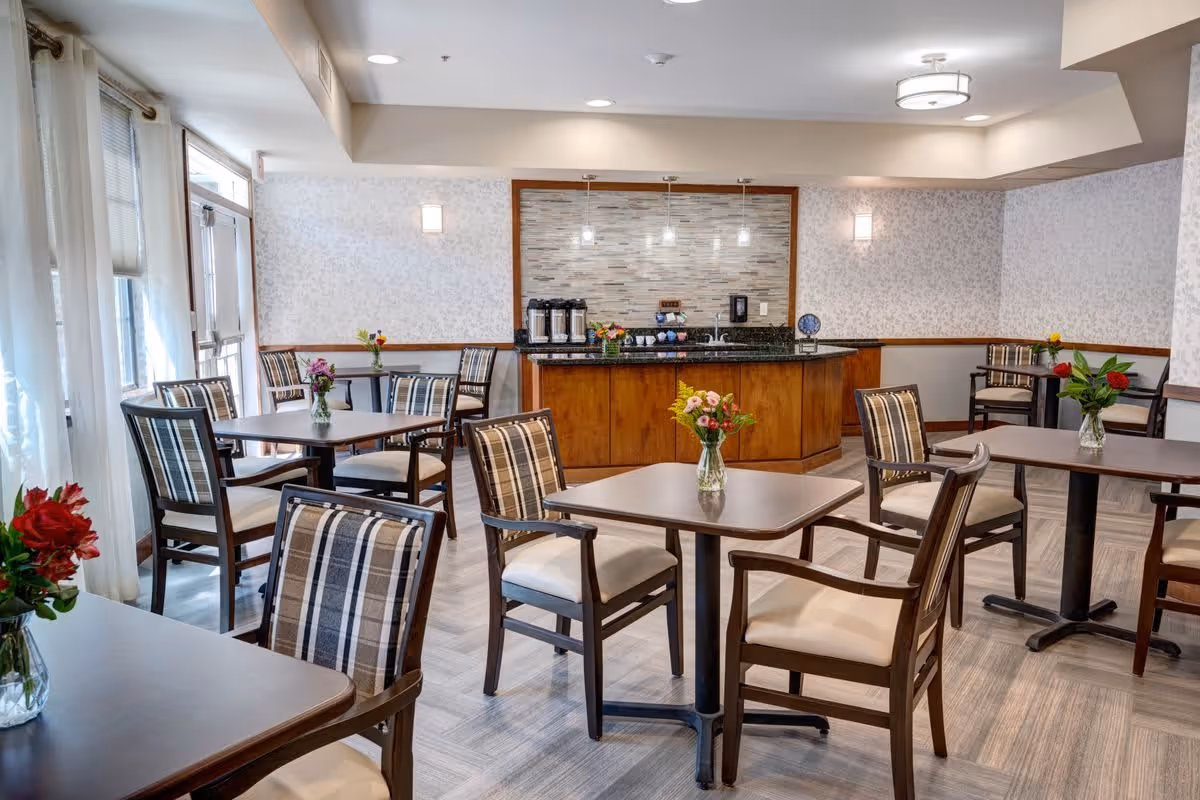 A bright dining room with several square tables and cushioned chairs arranged neatly. Each table has a small vase with colorful flowers. The room features large windows with sheer curtains allowing natural light to enter. At the back, there is a counter area with coffee dispensers and a tiled backsplash under soft ceiling lights.
