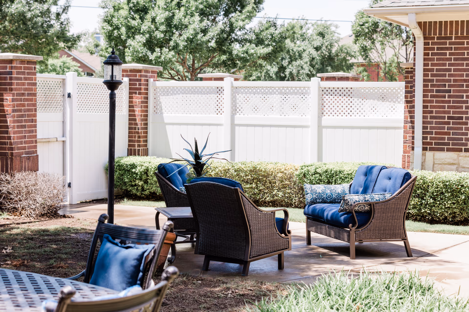 Outdoor patio area with cushioned wicker chairs and a loveseat arranged around a small table. The space is enclosed by a white fence with brick pillars, and there is a black lamp post near the seating area. Green shrubs and trees are visible in the background.