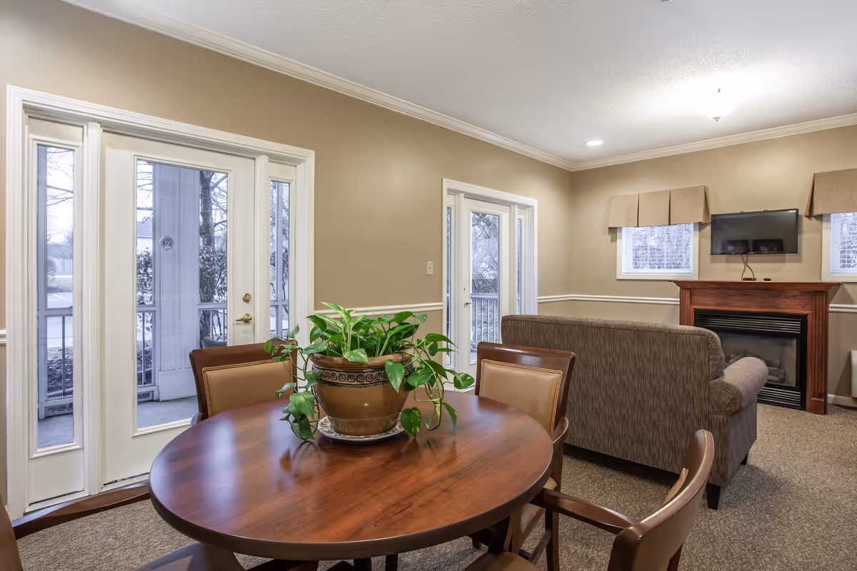 A cozy living room area with a round wooden table and four chairs in the foreground, featuring a potted green plant on the table. In the background, there is a brown upholstered sofa facing a fireplace with a mounted flat-screen TV above it. The room has beige walls, carpeted floor, and two sets of French doors leading outside, with windows on either side of the fireplace.