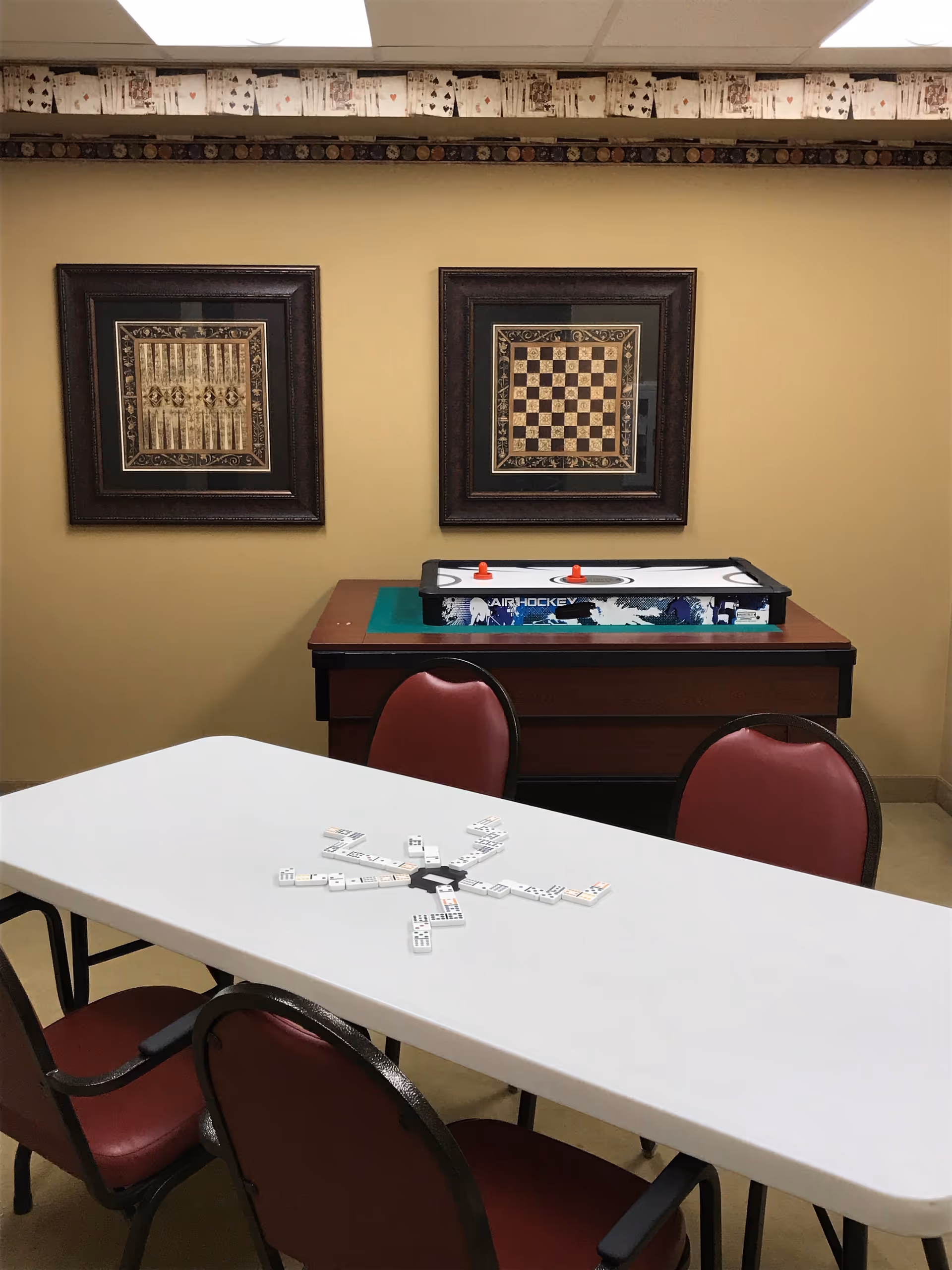 A game room with a white rectangular table surrounded by four red cushioned chairs. Dominoes are arranged in a circular pattern on the table. In the background, there is a small air hockey table with two red strikers on it. Two framed game boards, one for backgammon and one for chess, hang on a beige wall. A decorative border featuring playing cards runs along the top of the wall near the ceiling.