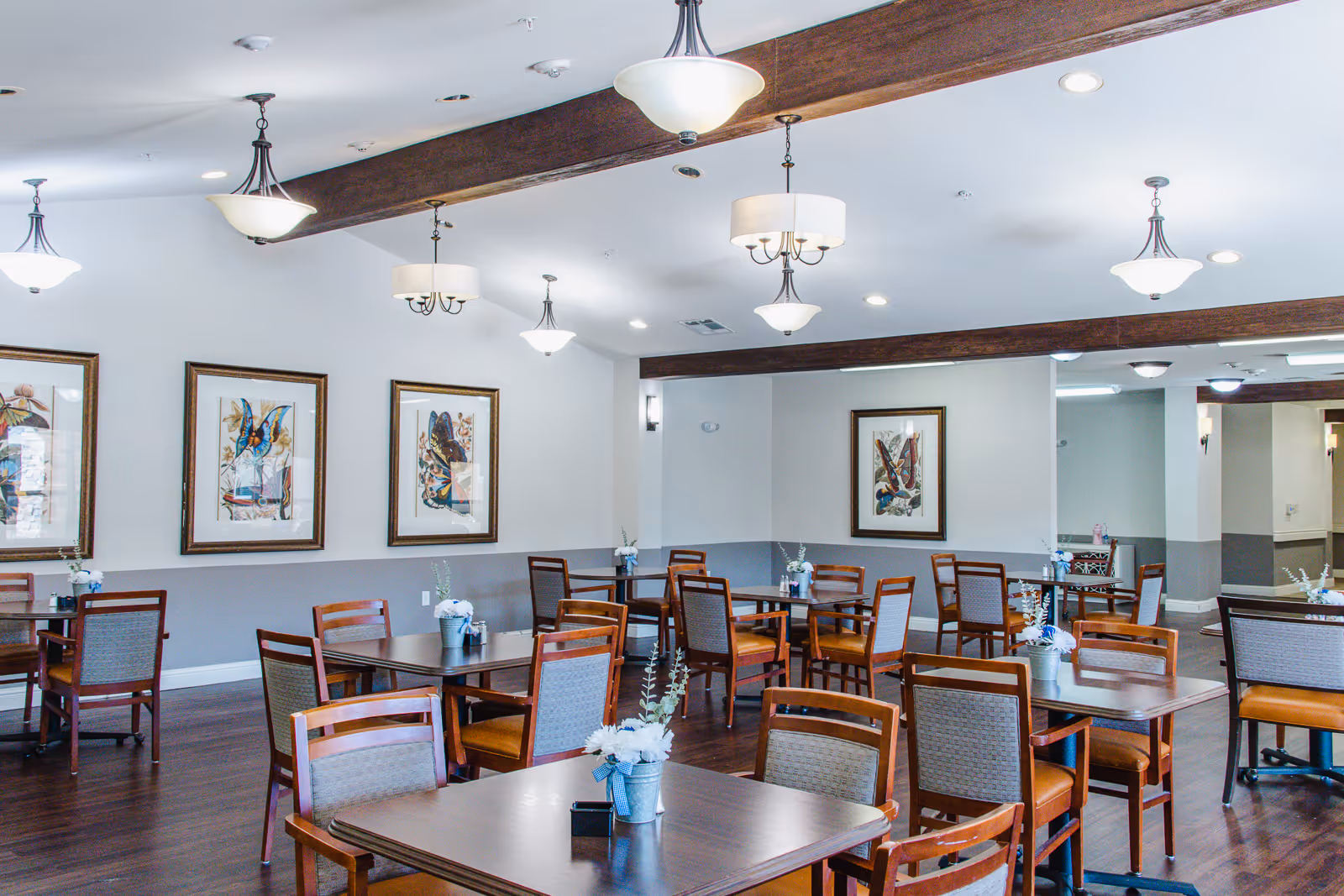Bright dining room with wooden tables and chairs, framed artwork on the walls, and multiple hanging light fixtures.