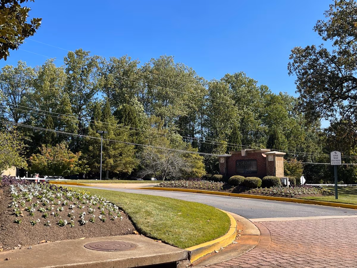Landscaped entrance with a curved driveway and a brick sign for St. George Village surrounded by shrubs and tall trees under a clear blue sky.