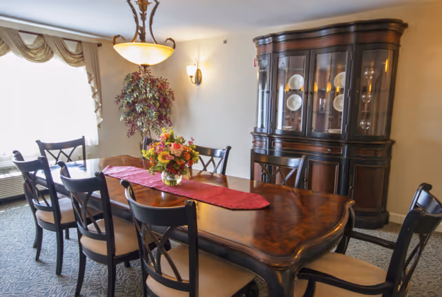 A formal dining room with a large wooden dining table surrounded by eight chairs with cushioned seats. A red table runner and a floral centerpiece decorate the table. In the background, there is a wooden china cabinet displaying plates and glassware. A large window with draped curtains allows natural light into the room, and a decorative ceiling light fixture hangs above the table.
