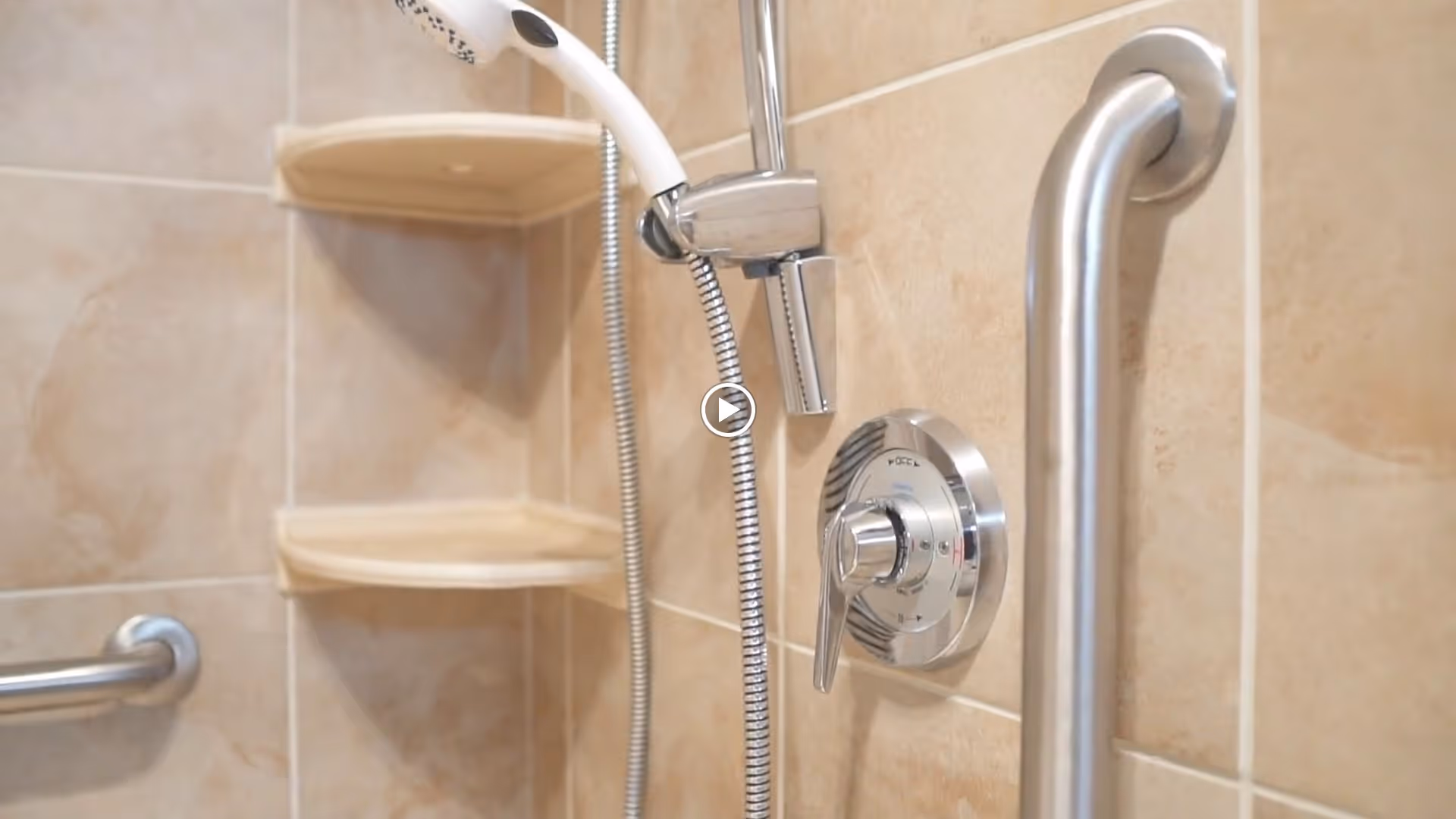 Close-up view of a shower area with beige tiled walls, featuring a handheld showerhead attached to a metal hose, a temperature control knob, and two built-in corner shelves. There are also two stainless steel grab bars installed on the walls for safety.