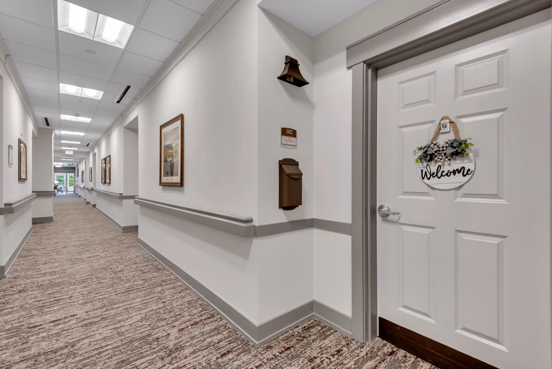 A clean, well-lit hallway in a senior living facility with beige walls, carpeted floor, and framed artwork on the walls. A white door with a 'Welcome' sign hangs on the right side, labeled room 102. The hallway extends into the distance with more doors and handrails along the walls.