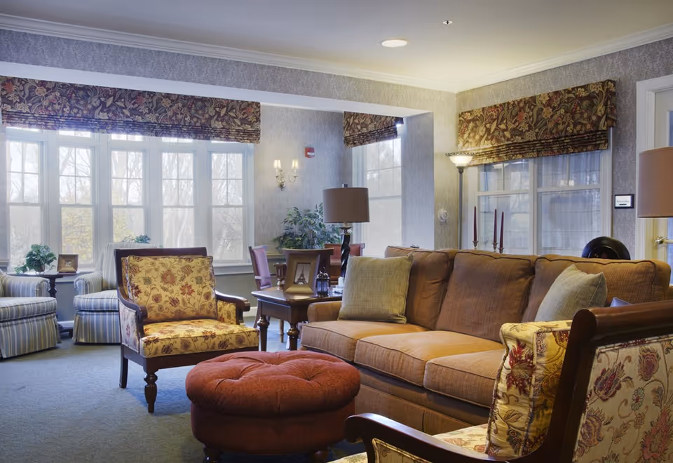 A cozy living room with a brown sofa, floral upholstered armchairs, a round red ottoman, and a wooden side table with a lamp. Large windows with floral valances allow natural light to fill the room. The walls have a subtle patterned wallpaper, and there are additional chairs and plants near the windows.